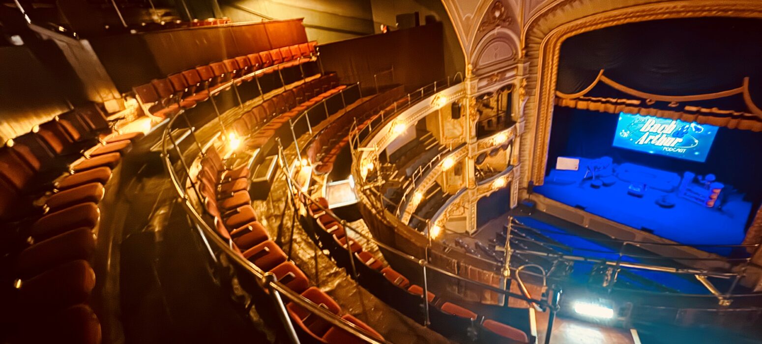 A photograph of of the Tyne Theatre & Opera House auditorium, taken from the Gallery. The photo shows the newly restored Gallery, its new red Heritage seating, and the stage which sits below on the right hand side of the image and is washed in blue lighting.