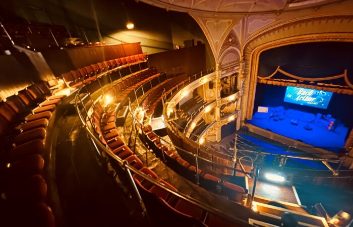 Gallery PIc – Credit Tyne Theatre & Opera House A photograph of of the Tyne Theatre & Opera House auditorium, taken from the Gallery. The photo shows the newly restored Gallery, its new red Heritage seating, and the stage which sits below on the right hand side of the image and is washed in blue lighting.