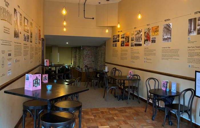 A photograph of the new Stoll Exhibition displayed on the walls of the 1867 Café at Tyne Theatre and Opera House. The exhibition spans the walls on either side of the cafe, displaying archival images and text, with QR codes for interactive elements.