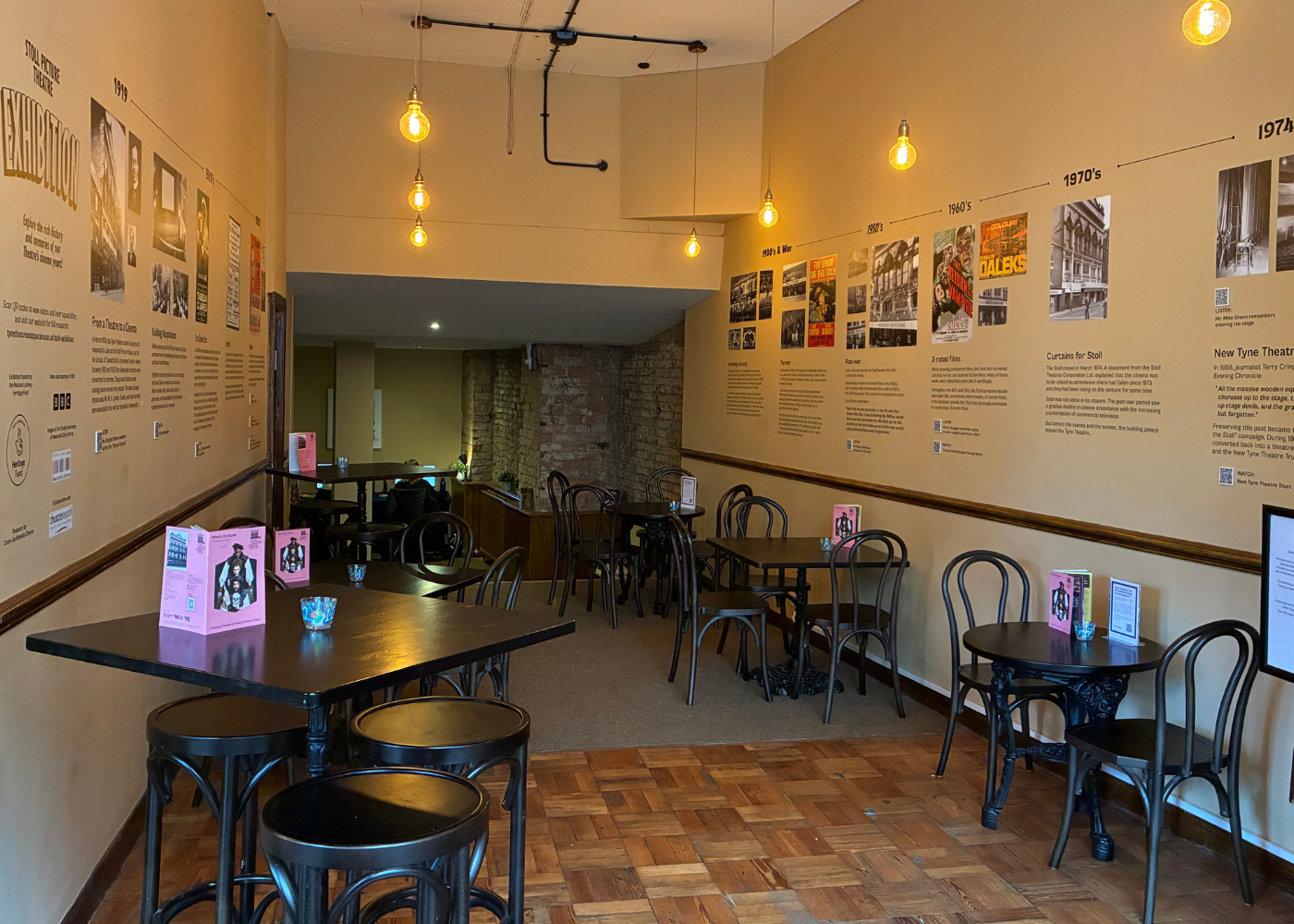 A photograph of the new Stoll Exhibition displayed on the walls of the 1867 Café at Tyne Theatre and Opera House. The exhibition spans the walls on either side of the cafe, displaying archival images and text, with QR codes for interactive elements.